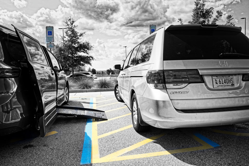 Picture is mostly in black and white with just the disabled hash marked space in color. A van is pulled up to about half of the hash marked space, and the ramp from our van is open, showing how there is no room for a wheelchair to get into our van.