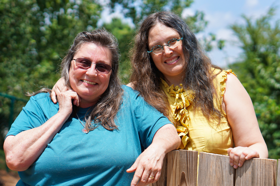 Tami stands on the left, and Veronica on the right. Veronica has her arm around Tami, and Tami is reaching up to hold Veronica's hand.