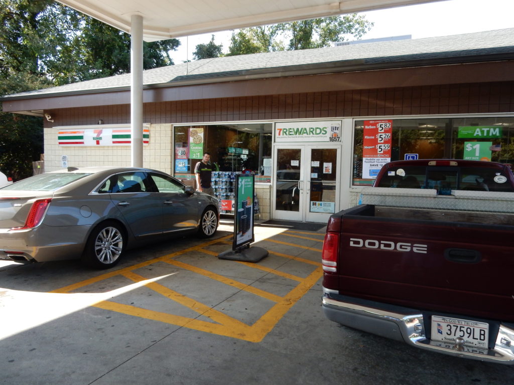 Outside the 7 eleven store, there is a yellow hash-marked space that is supposed to be available for people with disabilities to use to get out of their cars and get safely into the store. There is a sign selling soft drinks right in the middle of the walkway.