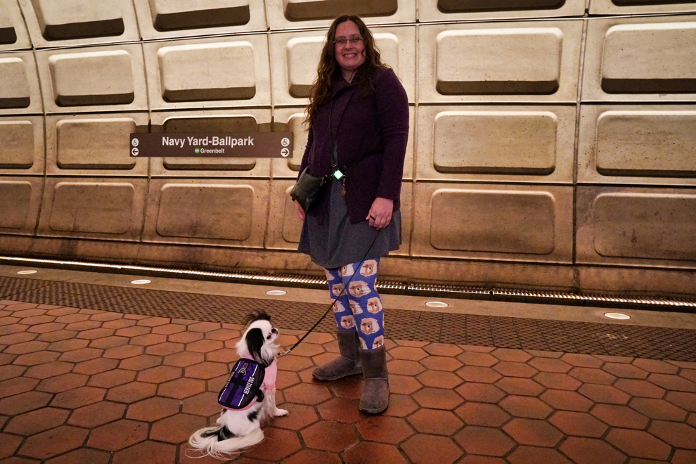 Veronica is wearing purple guinea pig leggings and is standing in the Metro station smiling at the camera. Hestia is sitting in front of Veronica, looking down the track.