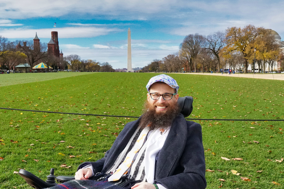 Brad is wearing a grey coat, a white blazer, a fox tie, a fox pin, and a blue and purple flower hat with lace on it. He poses in front of the Washington Monument far in the background.