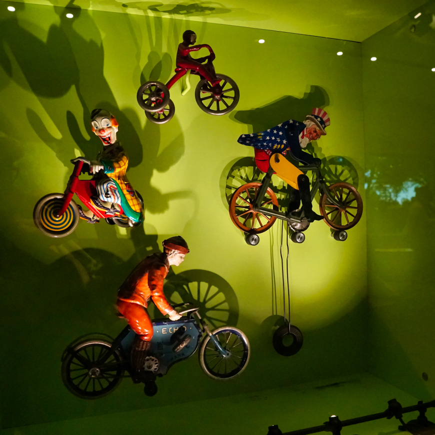 In front of a green background, several old bicyclist toys are displayed on a wall. One is a creepy clown, smiling while looking your way.