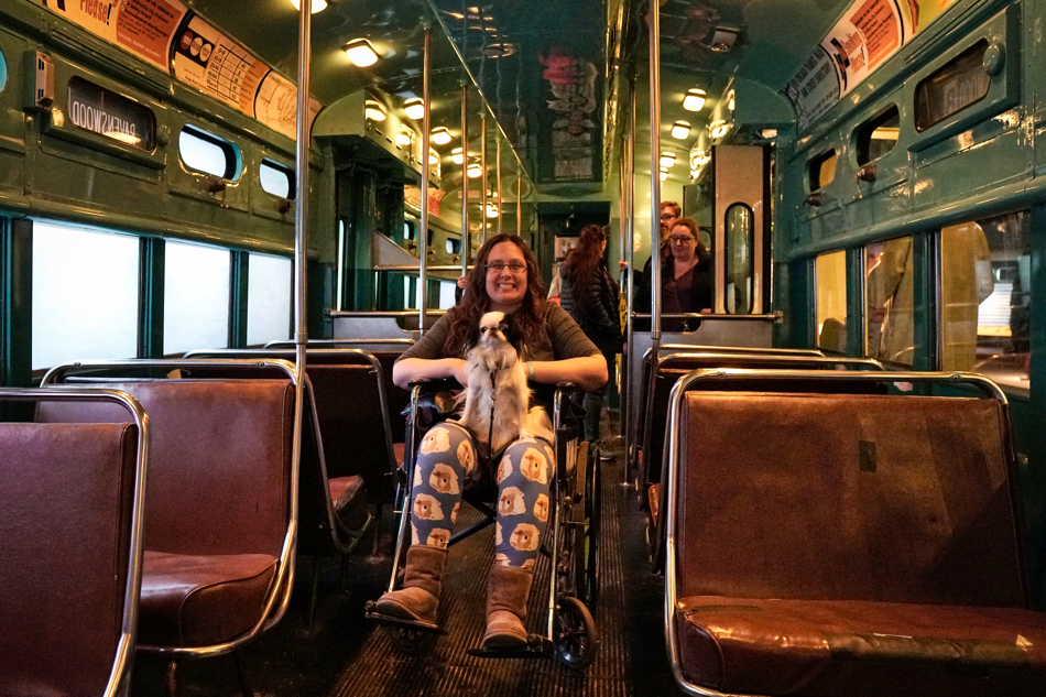 Veronica in a wheelchair in the aisle of a subway train in the museum. Hestia sits on her lap and they both smile at the camera.