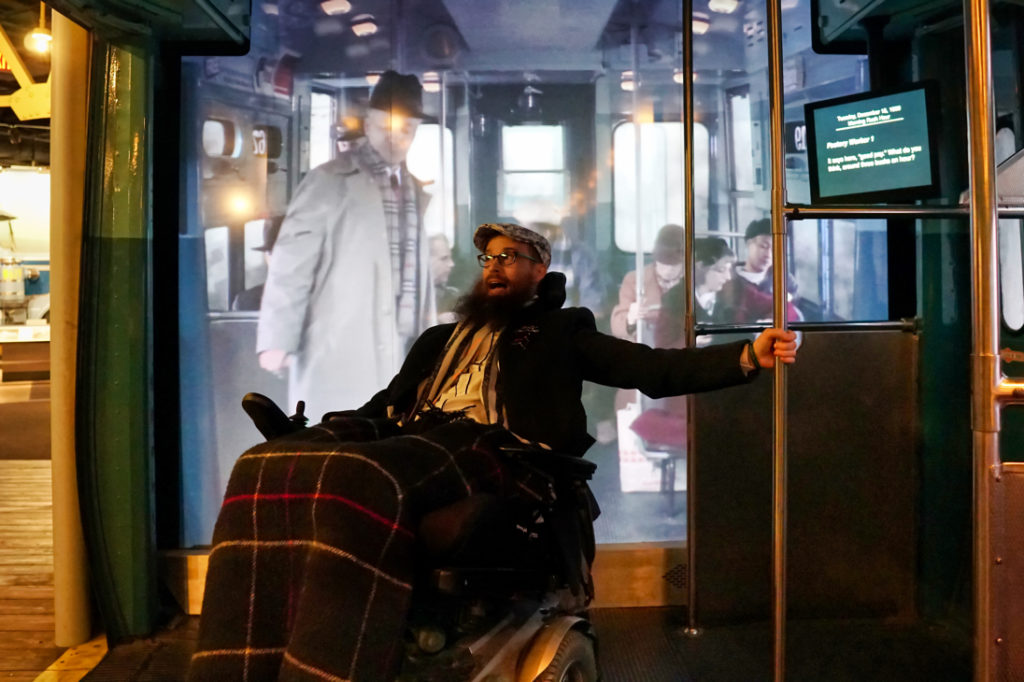 Brad holds onto a pole in the subway train as if he's sliding around, and in the background a movie is playing of what life on the subway would have been like in the 70s.