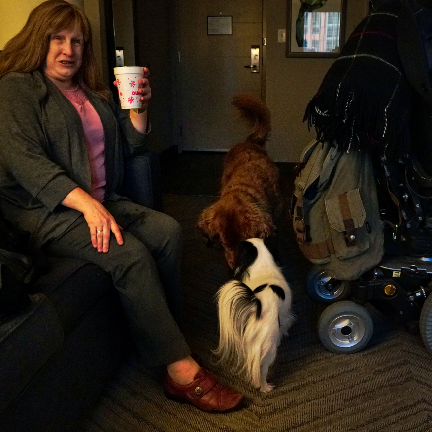 Jenine, a woman wearing a pink shirt and grey sweater and pants, sits on the couch while Roger and Hestia sniff each others' faces.
