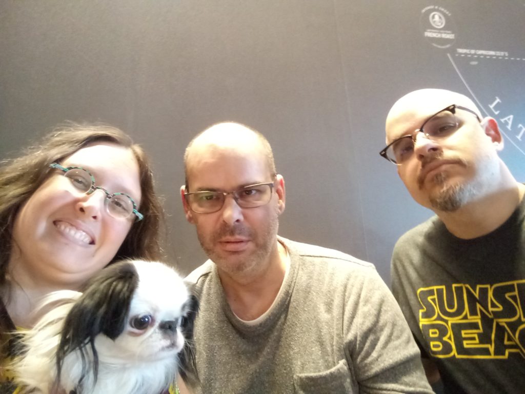 A selfie of us three! I am a woman with long brown curly hair on the left. On my lap is a black and white dog with a smushed nose. In the middle is Dan, who has on a grey shirt, has a bit of a beard, and is wearing glasses. Doc is on the right, wearing a Sunset Beach shirt, with a goatee and glasses. I am the only one smiling, everyone else is looking serious!